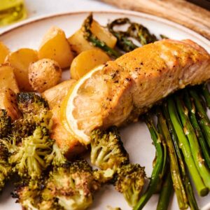 Sheet Pan Salmon baked to perfection with a lemon slice, served alongside roasted broccoli, green beans, and small potatoes.