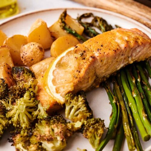 Sheet Pan Salmon baked to perfection with a lemon slice, served alongside roasted broccoli, green beans, and small potatoes.