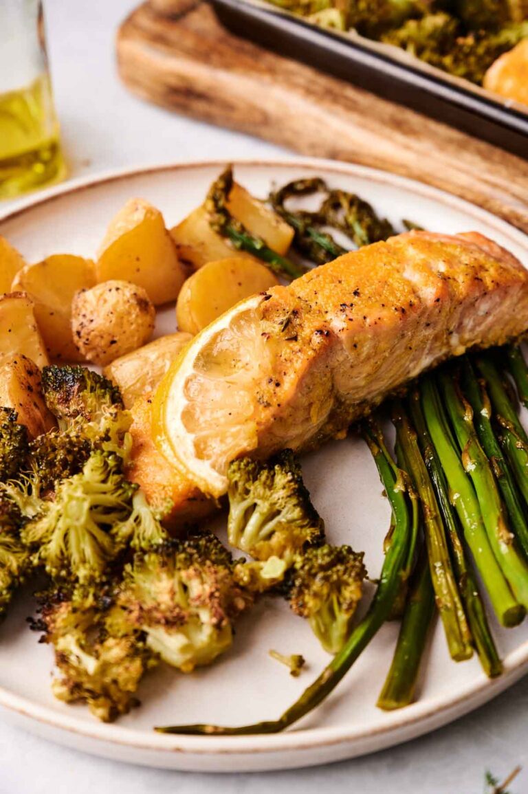 Sheet Pan Salmon baked to perfection with a lemon slice, served alongside roasted broccoli, green beans, and small potatoes.