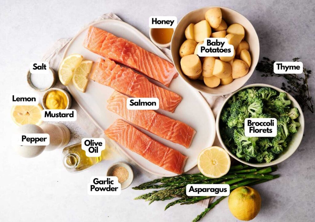 Top-down view of Sheet Pan Salmon fillets on a platter, surrounded by labeled ingredients: baby potatoes, broccoli, asparagus, lemon, mustard, garlic powder, olive oil, honey, salt, pepper, and thyme.