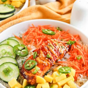 A bowl of rice topped with sliced grilled salmon, shredded carrots, cucumber, mango cubes, jalapeño slices, green onions, and sesame seeds—these Spicy Salmon Bowls are a fresh and flavorful meal.