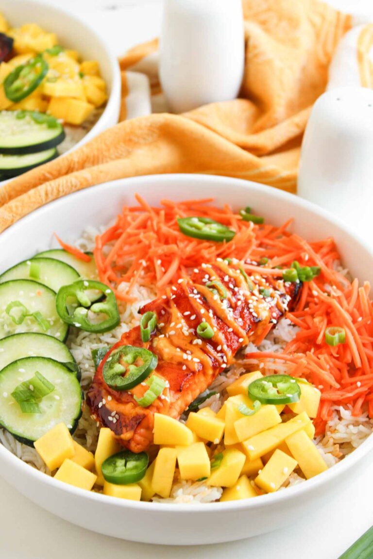 A bowl of rice topped with sliced grilled salmon, shredded carrots, cucumber, mango cubes, jalapeño slices, green onions, and sesame seeds—these Spicy Salmon Bowls are a fresh and flavorful meal.