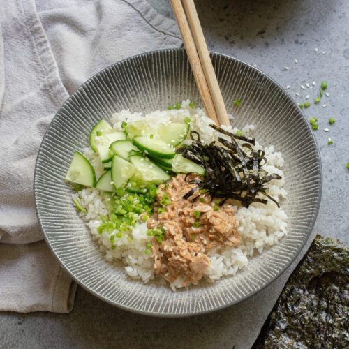 A Spicy Tuna Bowl featuring white rice topped with sliced cucumber, shredded seaweed, chopped green onions, and a creamy spicy tuna mixture, with chopsticks resting on the bowl.