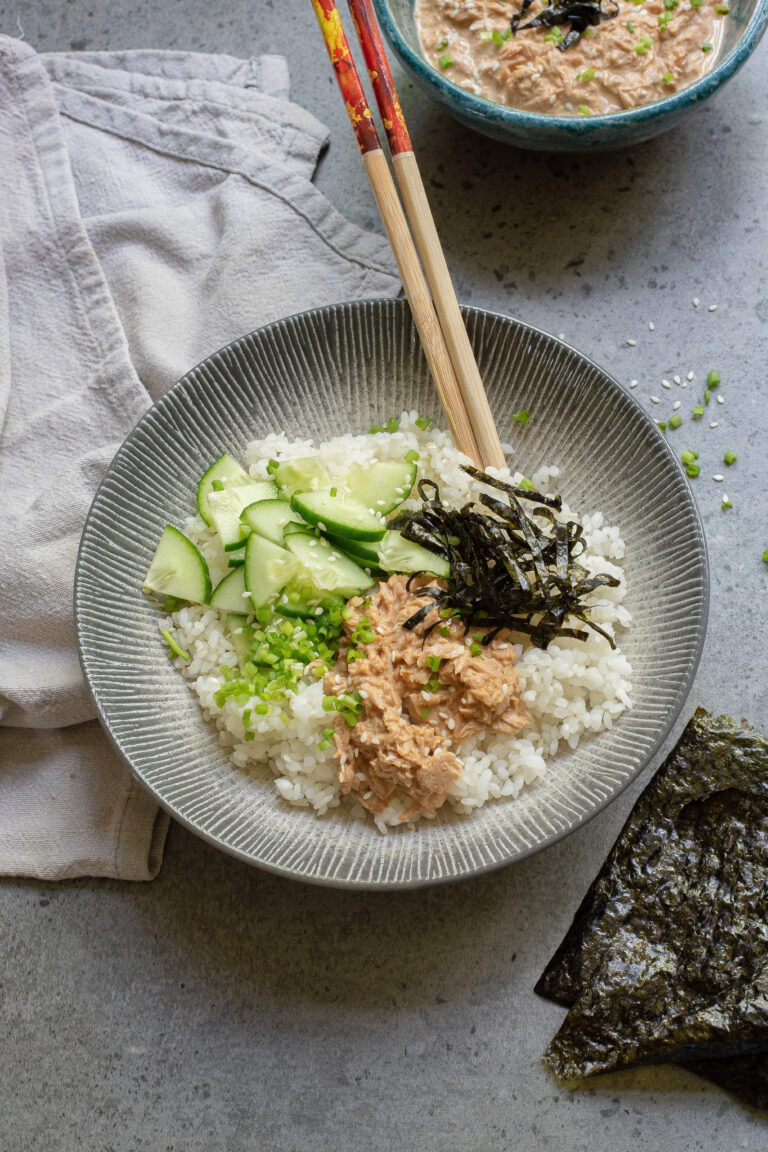 A Spicy Tuna Bowl featuring white rice topped with sliced cucumber, shredded seaweed, chopped green onions, and a creamy spicy tuna mixture, with chopsticks resting on the bowl.