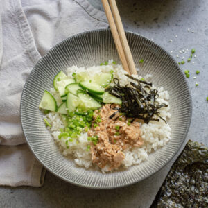 A Spicy Tuna Bowl featuring white rice topped with sliced cucumber, shredded seaweed, chopped green onions, and a creamy spicy tuna mixture, with chopsticks resting on the bowl.
