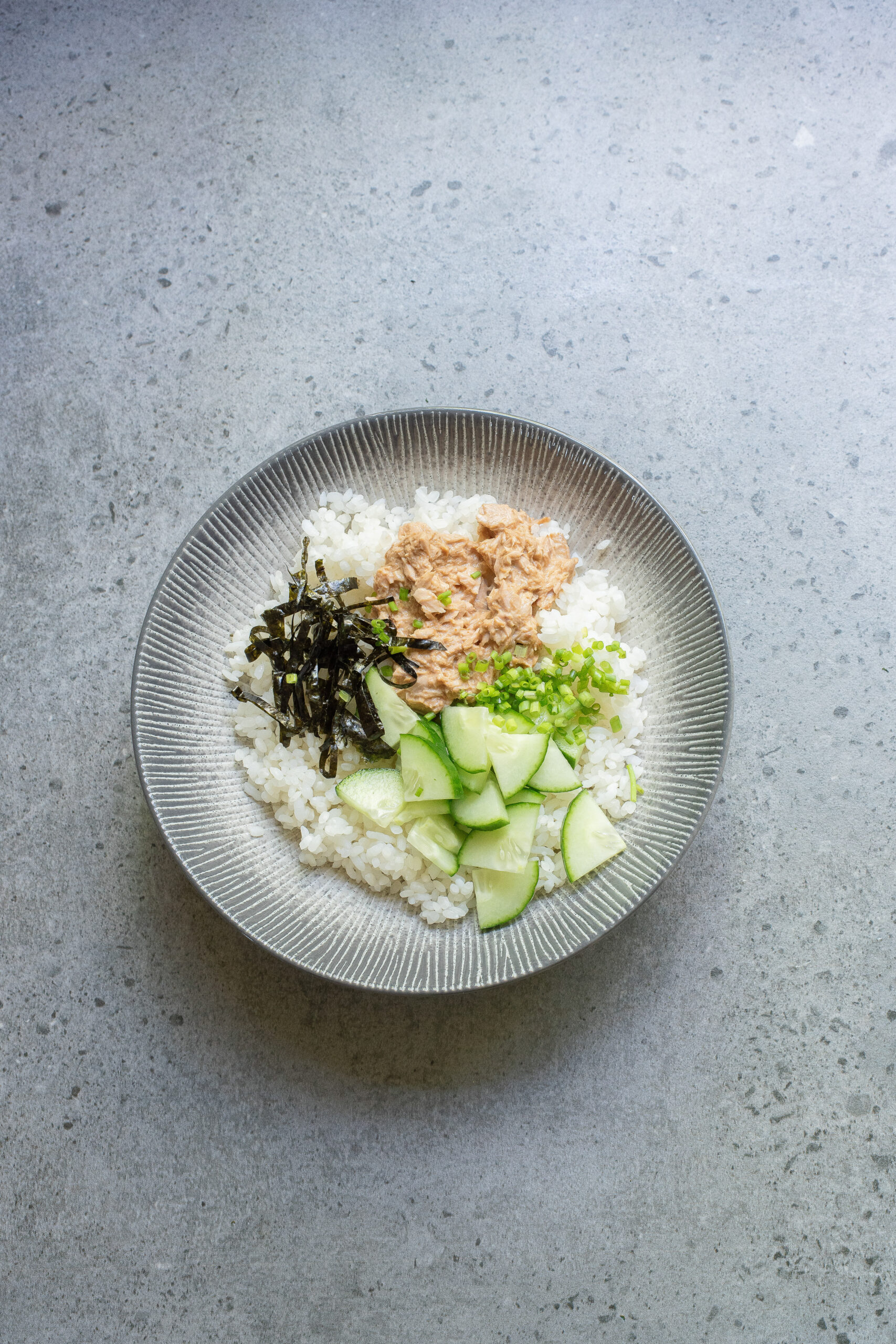 A Spicy Tuna Bowl featuring a bed of white rice topped with shredded seaweed, tuna, sliced cucumber, and chopped green onions on a grey surface.