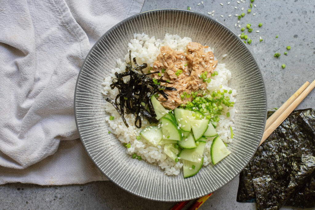 A Spicy Tuna Bowl featuring a bed of white rice topped with cucumber slices, seaweed strips, a flavorful tuna mixture, and chopped green onions, served with chopsticks and extra nori sheets on the side.