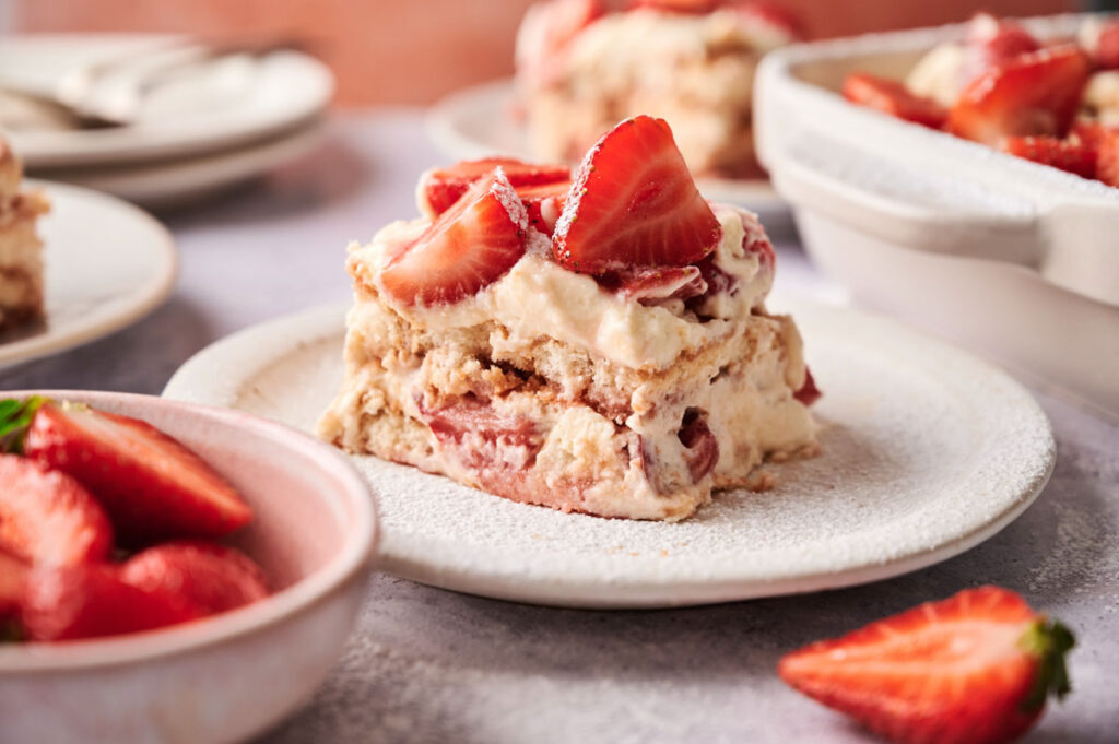 A slice of Strawberry Tiramisu topped with fresh strawberry slices sits on a white plate, with a bowl of strawberries and more Strawberry Tiramisu in the background.