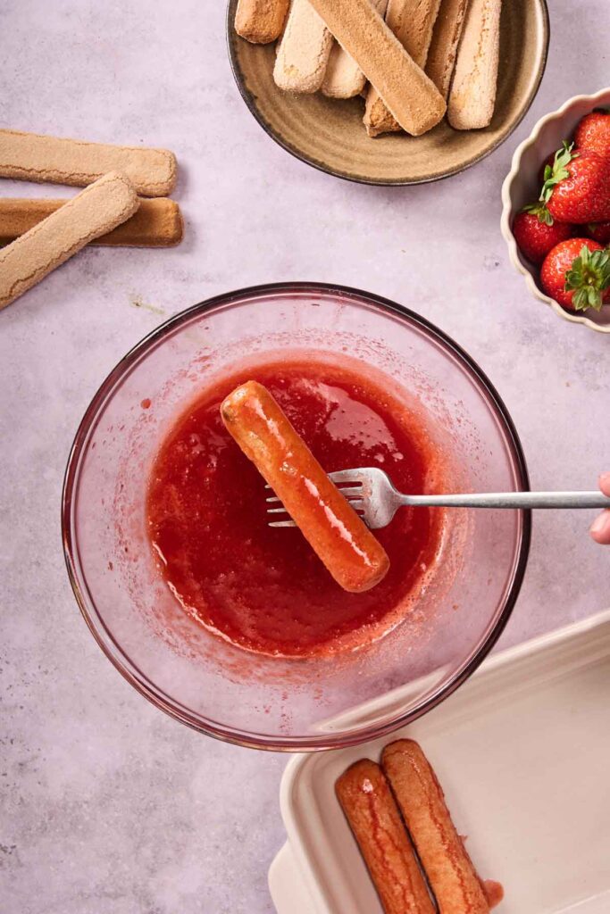 A hand dips a ladyfinger biscuit into a bowl of strawberry sauce—perfect for assembling a Strawberry Tiramisu; more ladyfingers and strawberries rest on the counter nearby.
