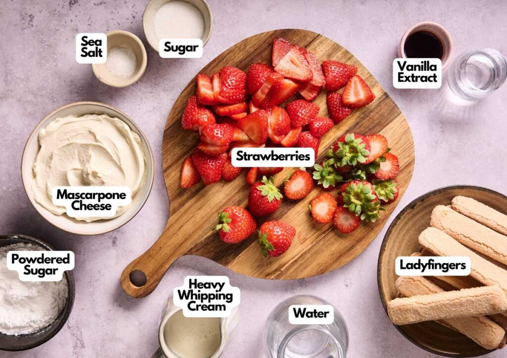 A round wooden board with sliced and whole strawberries, surrounded by labeled bowls of mascarpone cheese, heavy whipping cream, powdered sugar, sugar, sea salt, vanilla extract, water, and ladyfingers for Strawberry Tiramisu.