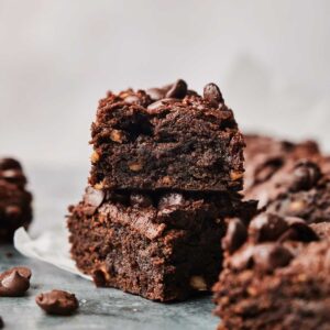 Two stacked Sweet Potato Brownies with chocolate chips on top, surrounded by more brownies and scattered chocolate chips on a dark surface.