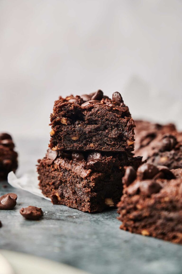 Two stacked Sweet Potato Brownies with chocolate chips on top, surrounded by more brownies and scattered chocolate chips on a dark surface.