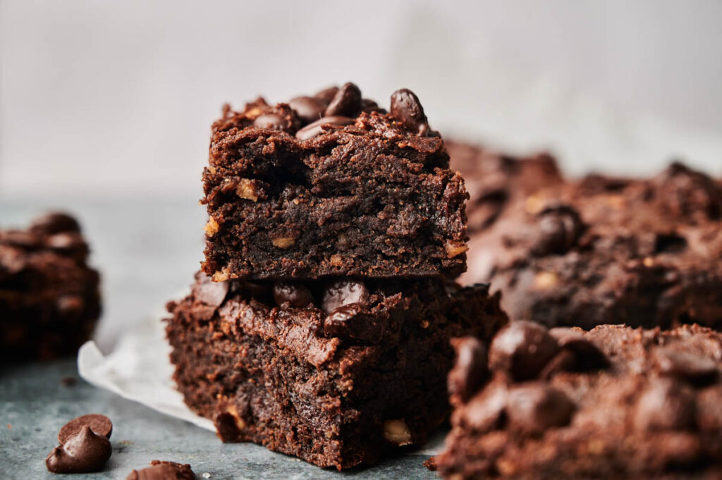 Two sweet potato brownies stacked on top of each other, topped with chocolate chips, with more brownies in the background.