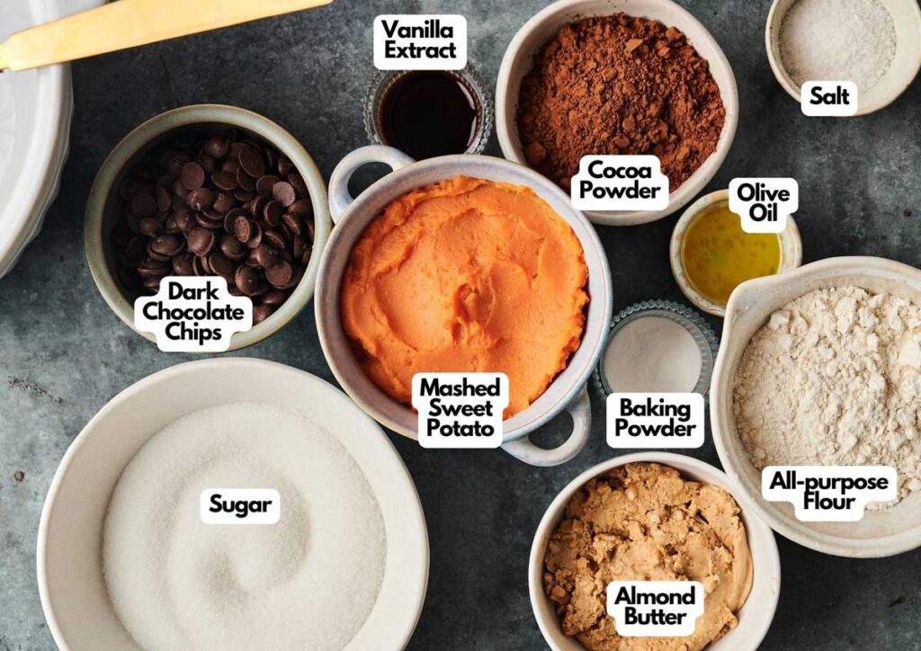 Bowls of ingredients for baking Sweet Potato Brownies, including mashed sweet potato, sugar, almond butter, all-purpose flour, cocoa powder, dark chocolate chips, olive oil, vanilla extract, baking powder, and salt.