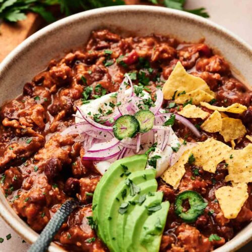 A bowl of Turkey Chili topped with sliced avocado, red onion, jalapeño, sour cream, cilantro, and tortilla chips, with a garnish of fresh parsley in the background.