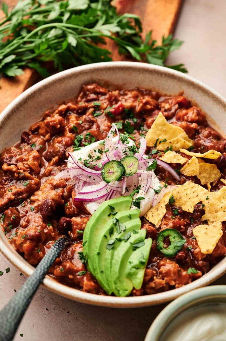 A bowl of Turkey Chili topped with sliced avocado, red onion, jalapeño, sour cream, cilantro, and tortilla chips, with a garnish of fresh parsley in the background.