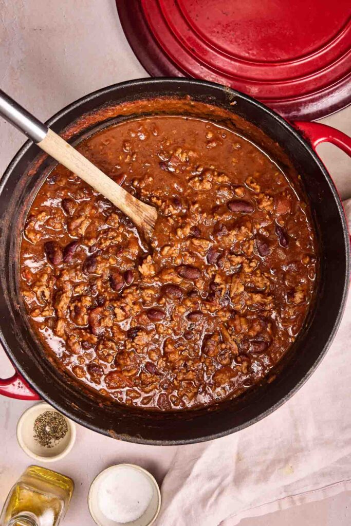 A pot of Turkey Chili with ground meat and kidney beans is stirred with a wooden spoon. Seasonings and oil rest nearby on a white cloth.