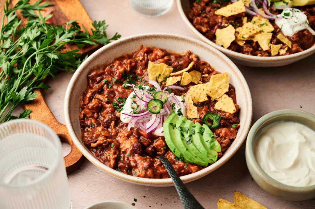 A bowl of hearty turkey chili topped with avocado slices, red onion, jalapeño, tortilla chips, and herbs, served with a side of sour cream and fresh parsley.