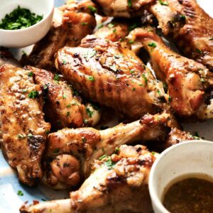 A plate of roasted chicken drumsticks, thighs, and turkey wings garnished with herbs, served with bowls of chopped parsley and dipping sauce.