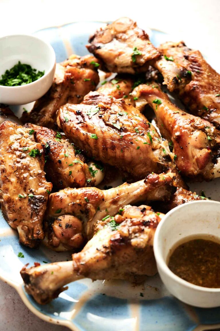 A plate of roasted chicken drumsticks, thighs, and turkey wings garnished with herbs, served with bowls of chopped parsley and dipping sauce.