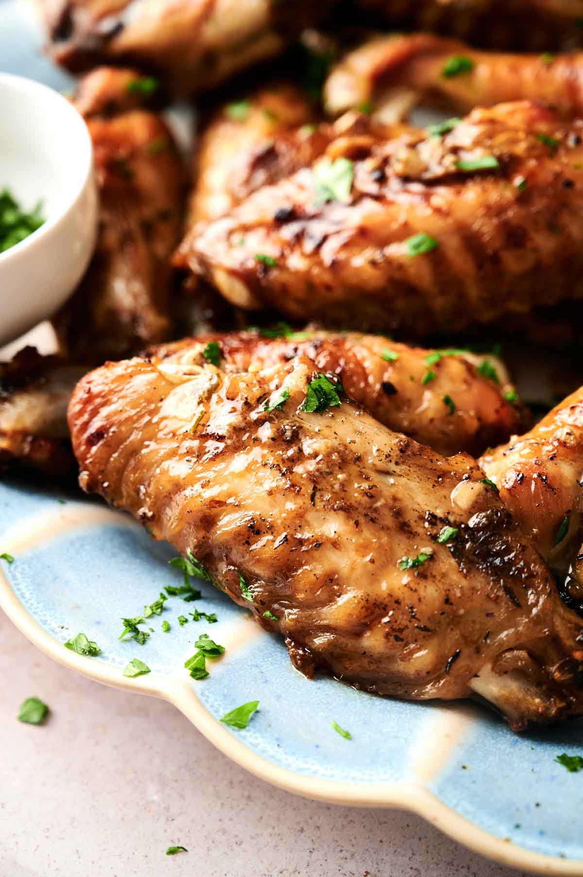Close-up of grilled chicken and turkey wings garnished with chopped parsley on a blue plate, with a small bowl of sauce in the background.