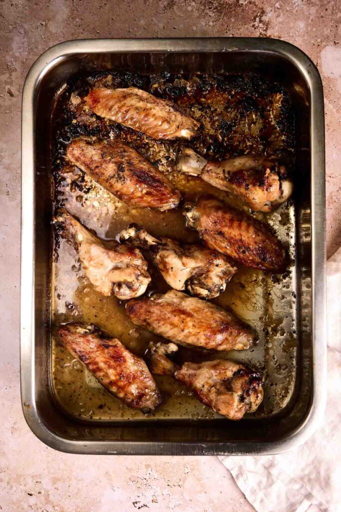 A metal baking tray with nine cooked chicken and turkey wings in oil and seasonings on a beige surface.