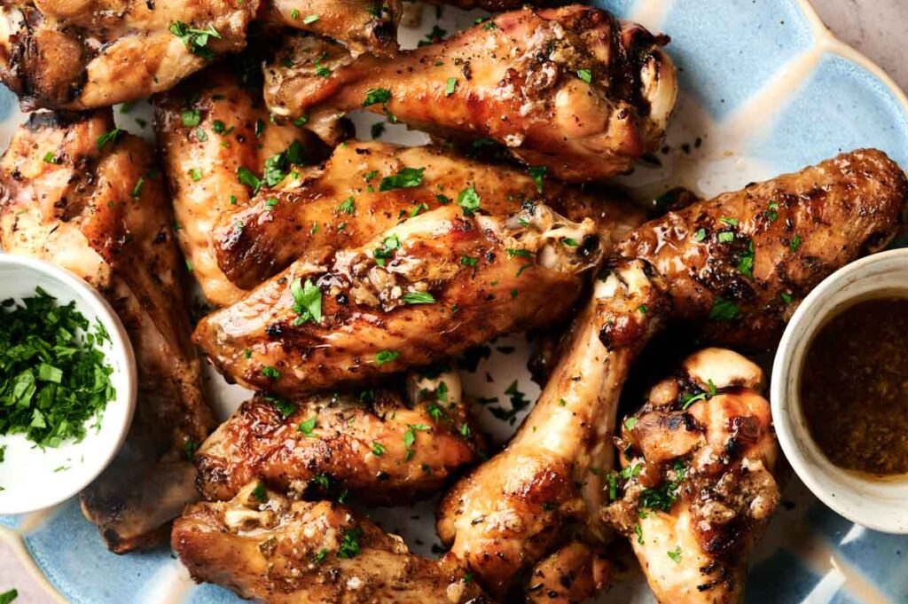 A plate of roasted chicken and turkey wings garnished with chopped herbs, accompanied by two small bowls containing dipping sauce and fresh herbs.