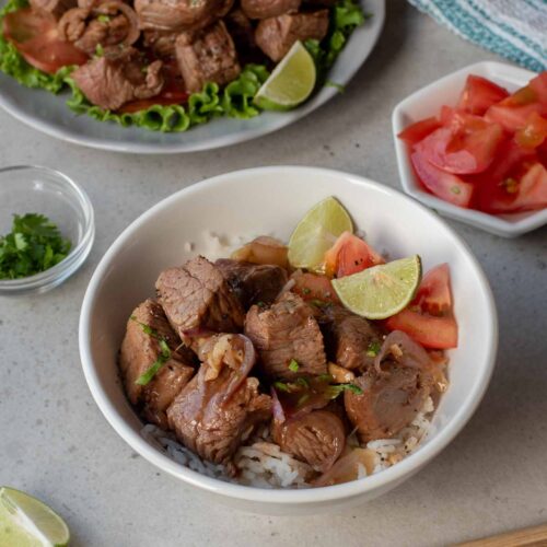 A bowl of white rice topped with Vietnamese Shaking Beef cubes, tomato slices, and lime wedges, with chopsticks, extra beef, lime, cilantro, and tomatoes on the side.