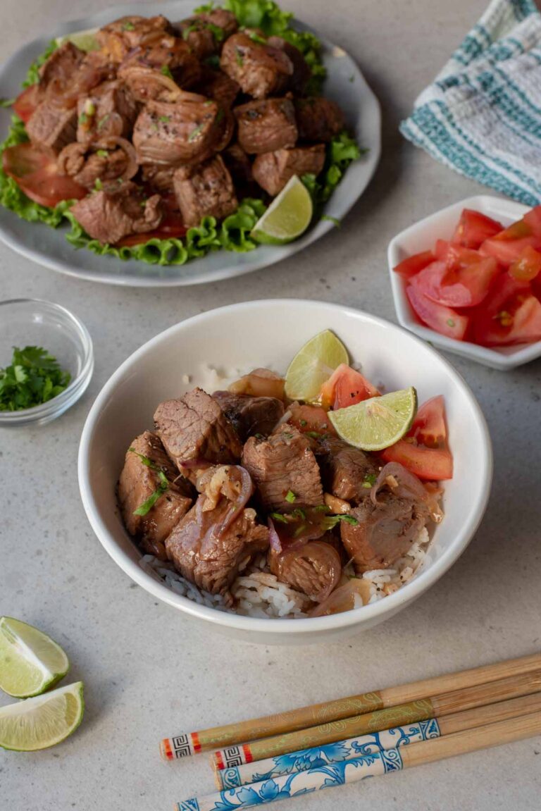 A bowl of white rice topped with Vietnamese Shaking Beef cubes, tomato slices, and lime wedges, with chopsticks, extra beef, lime, cilantro, and tomatoes on the side.