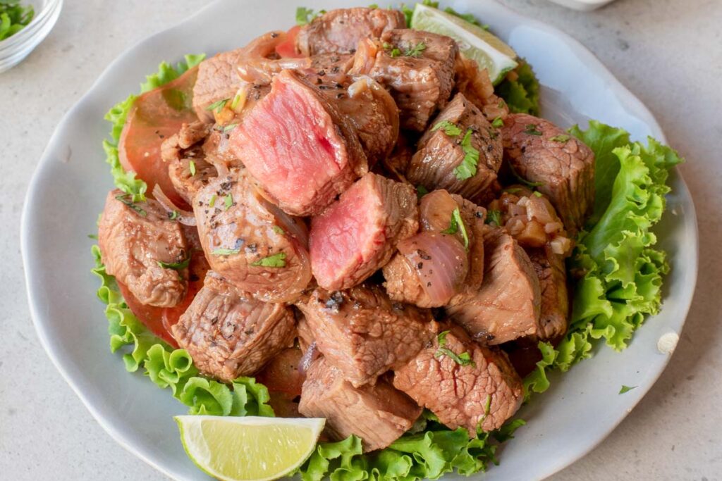 A plate of Vietnamese Shaking Beef features seared beef cubes garnished with chopped herbs, placed on a bed of lettuce with lime wedges on the side.