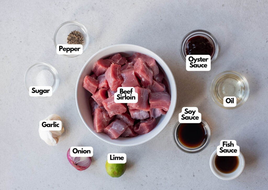 Bowl of raw beef sirloin for Vietnamese Shaking Beef, surrounded by small bowls of pepper, oyster sauce, oil, soy sauce, fish sauce, sugar, garlic, onion, and a lime on a light surface.