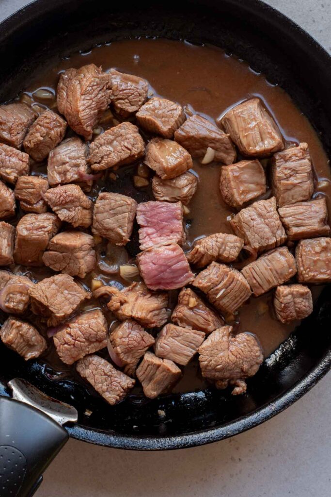 Cubed beef pieces are cooking in a black skillet with sauce, as in Vietnamese Shaking Beef, with one piece in the center noticeably less cooked than the others.
