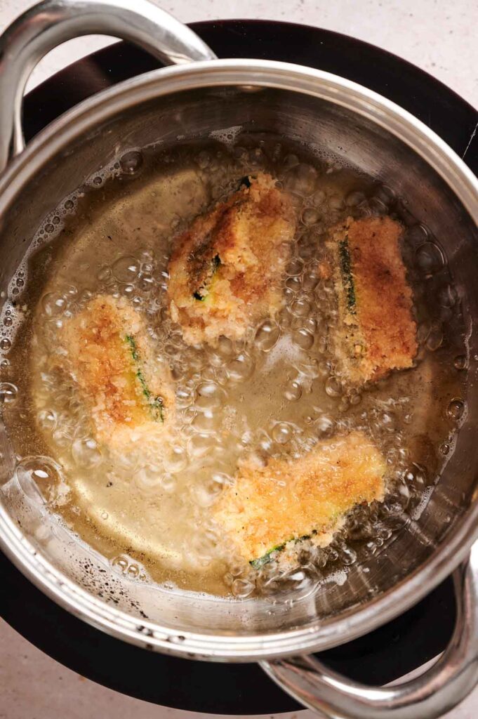 Four breaded zucchini fries are being deep-fried in hot oil inside a stainless steel pot on a stovetop.