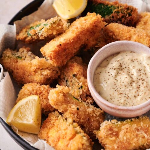 A bowl of crispy zucchini fries, breaded and fried to perfection, served with lemon wedges and a small bowl of creamy dipping sauce on paper towels.