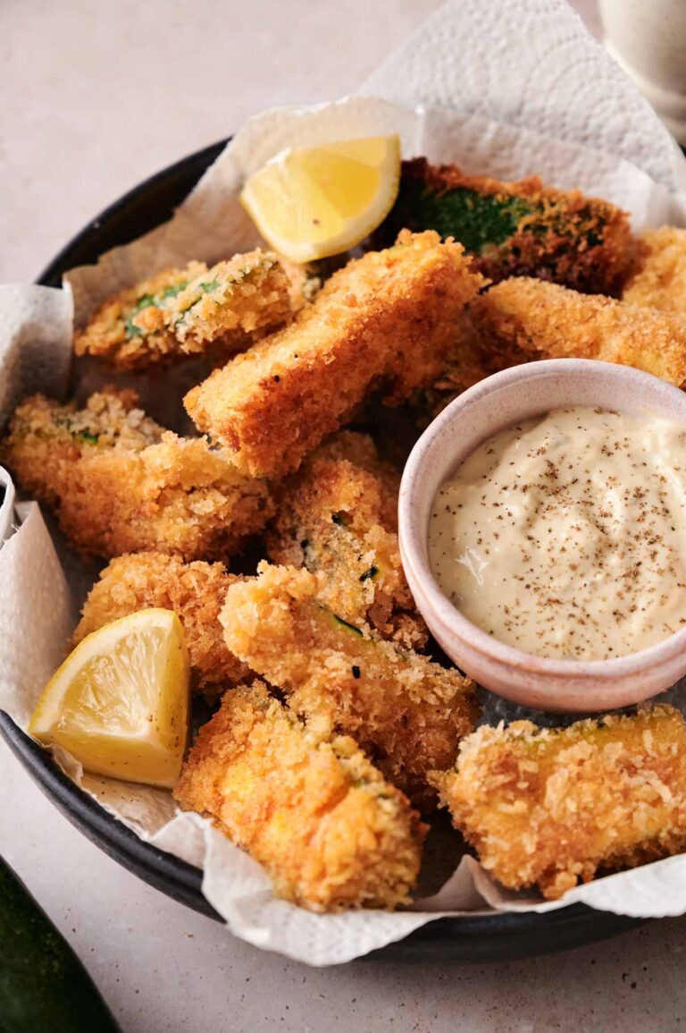 A bowl of crispy zucchini fries, breaded and fried to perfection, served with lemon wedges and a small bowl of creamy dipping sauce on paper towels.