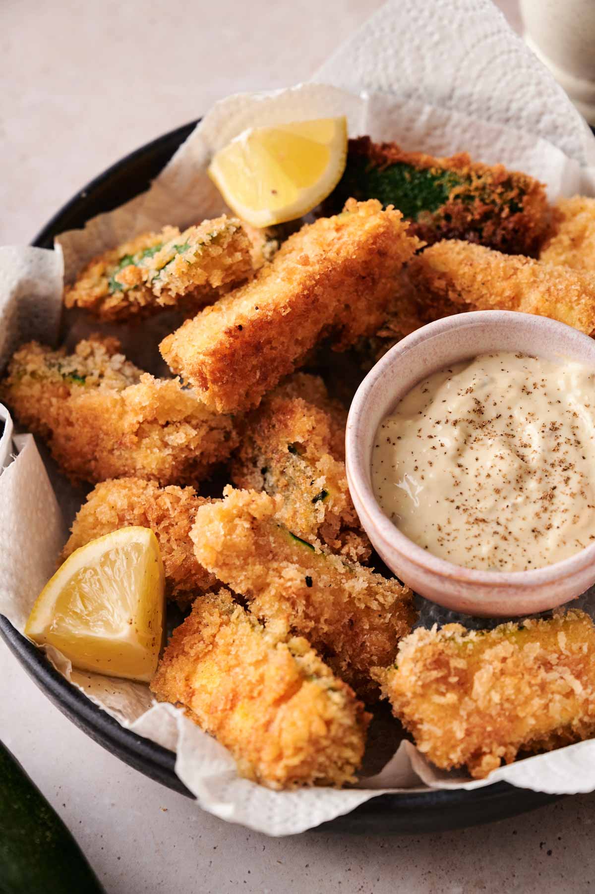 A bowl of crispy zucchini fries, breaded and fried to perfection, served with lemon wedges and a small bowl of creamy dipping sauce on paper towels.