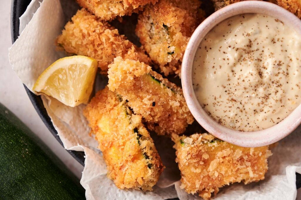 A bowl of crispy Zucchini Fries, breaded and fried to golden perfection, served with a wedge of lemon and a small bowl of creamy dipping sauce topped with black pepper.
