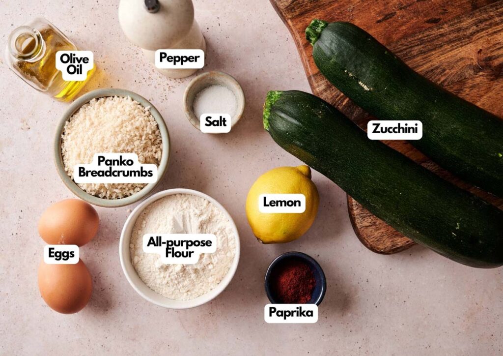 Various labeled ingredients for Zucchini Fries are arranged on a table, including zucchini, lemon, eggs, panko breadcrumbs, all-purpose flour, paprika, salt, pepper, and olive oil.