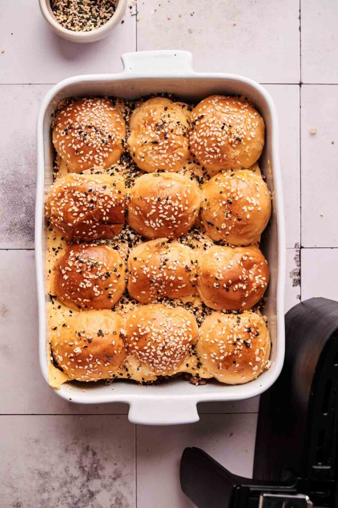 A white baking dish filled with twelve golden brown, sesame seed-topped Air Fryer Hamburger Sliders, partially cut and baked together. A small bowl of extra seeds sits nearby.