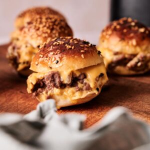 Three cheeseburgers with sesame seed buns and melted cheese are arranged on a wooden surface, perfect for serving as Air Fryer Hamburger Sliders at your next gathering.
