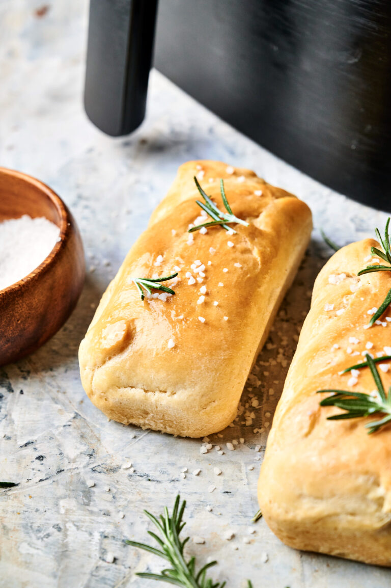 Two rectangular loaves of Air Fryer Sourdough Focaccia, topped with coarse salt and rosemary, rest on a light surface beside a small wooden bowl of salt.