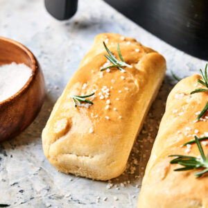 Two rectangular loaves of Air Fryer Sourdough Focaccia, topped with coarse salt and rosemary, rest on a light surface beside a small wooden bowl of salt.