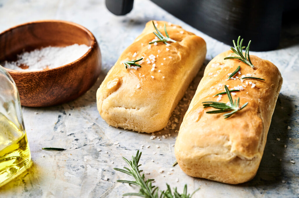 Two rectangular Air Fryer Sourdough Focaccia loaves, topped with rosemary and coarse salt, rest on a textured surface beside a bowl of salt and a bottle of olive oil.