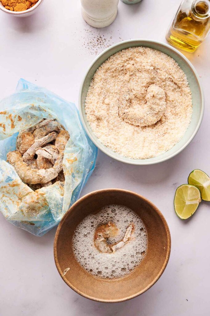 Bowls with breadcrumb coating, egg wash, and a plastic bag of raw shrimp are arranged on a white surface for Coconut Shrimp, with lime wedges and olive oil nearby.