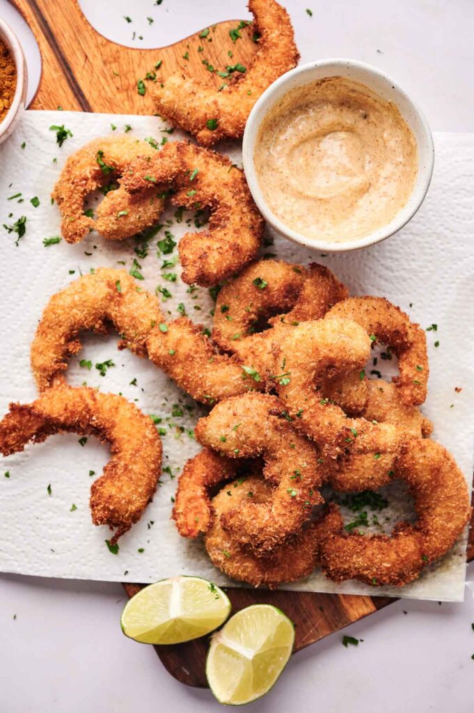 Crispy coconut shrimp are arranged on a paper towel with a bowl of dipping sauce, lemon wedges, and a sprinkle of chopped parsley.