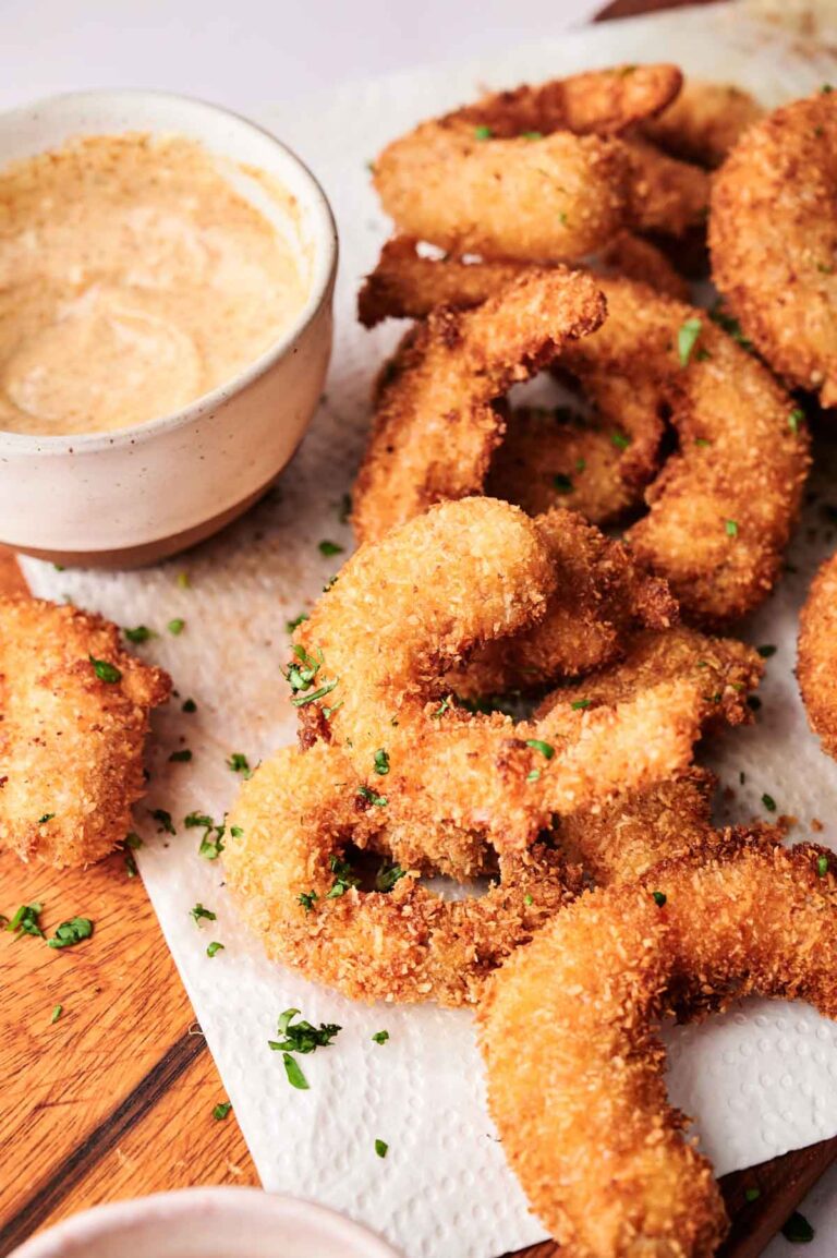 Crispy coconut shrimp, breaded and fried to golden perfection, arranged on a paper towel with a bowl of dipping sauce on a wooden surface.