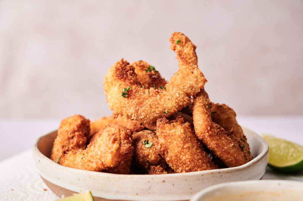 A ceramic bowl filled with golden-brown coconut shrimp, garnished with herbs, with a slice of lime and dipping sauce visible in the foreground.