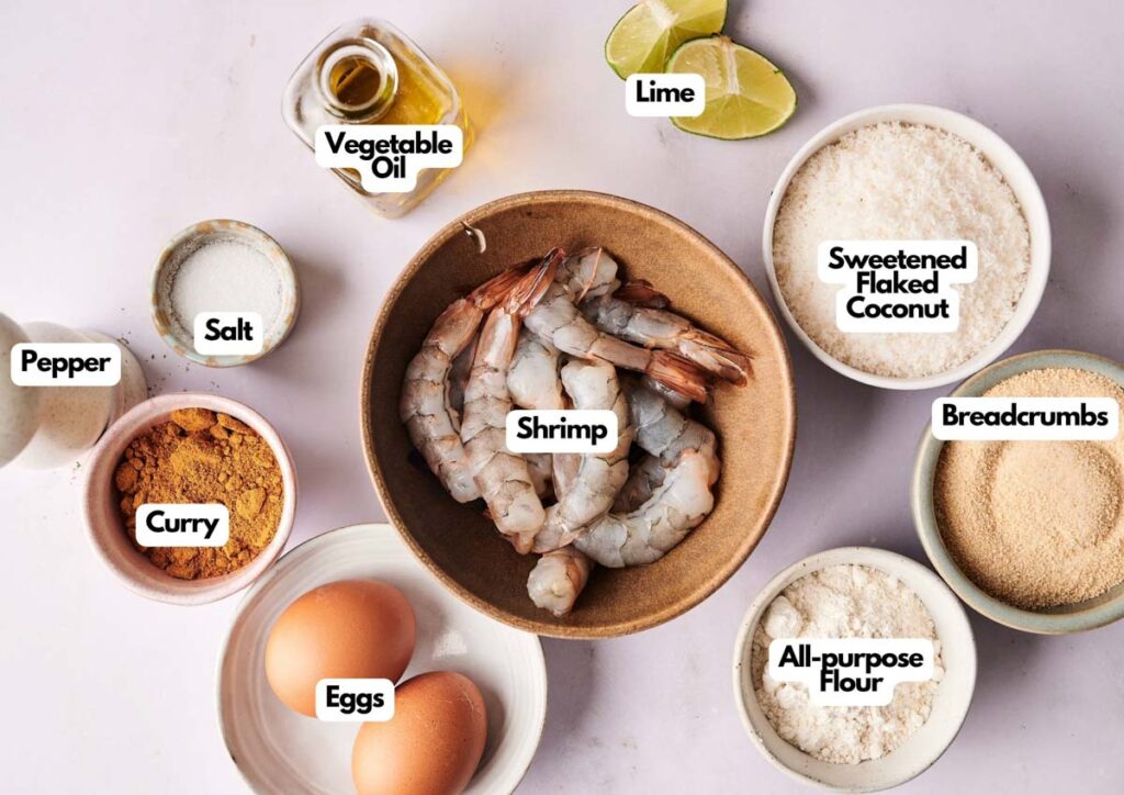 A bowl of raw shrimp, ready to become Coconut Shrimp, is surrounded by labeled ingredients: vegetable oil, lime, sweetened flaked coconut, breadcrumbs, all-purpose flour, eggs, curry powder, salt, and pepper.