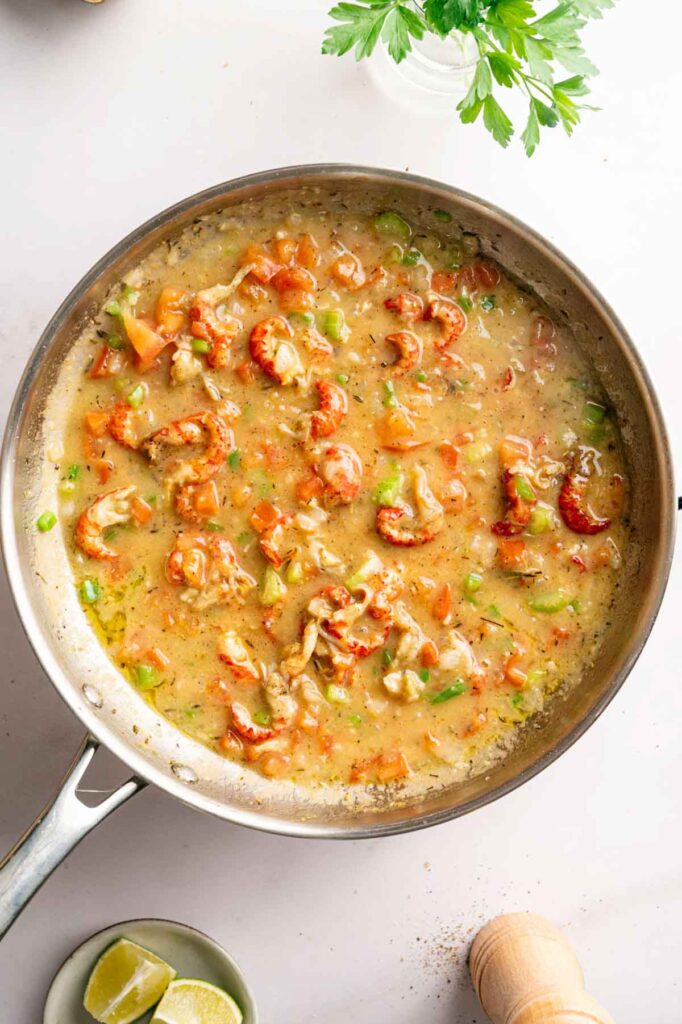 A skillet filled with a cooked mixture of crawfish tails, diced tomatoes, green peppers, and a light sauce evokes classic Crawfish Etouffee; lime wedges and parsley sit nearby on the white surface.