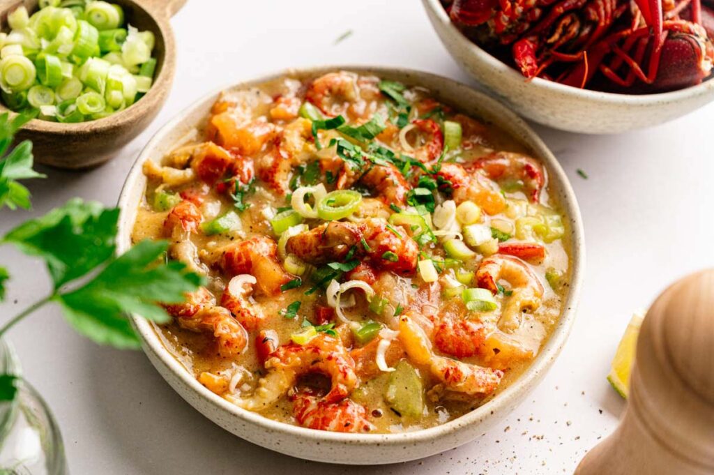A bowl of Crawfish Etouffee garnished with chopped green onions and parsley, with bowls of sliced green onions and whole crawfish nearby.